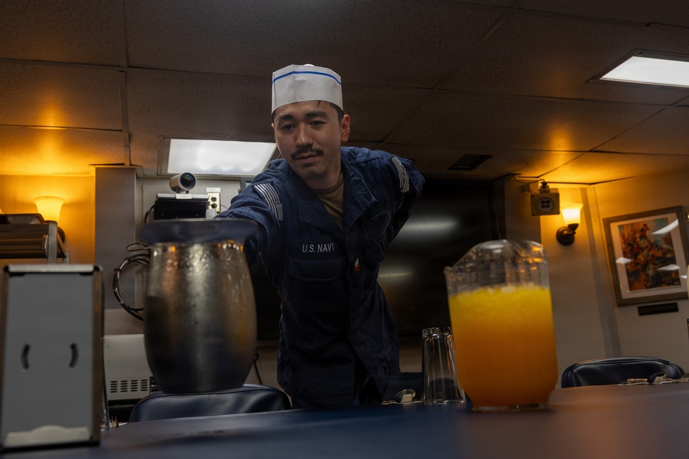 A U.S. Navy sailor wearing a white garrison cap and navy blue uniform is setting a table or serving area aboard what appears to be a naval vessel's wardroom or mess. The sailor is leaning forward over a table that holds beverage containers, including a large pitcher of orange juice and a metal pitcher. The warm ambient lighting and framed artwork on the wall suggest this is an officer's dining area or mess hall.