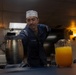 A U.S. Navy sailor wearing a white garrison cap and navy blue uniform is setting a table or serving area aboard what appears to be a naval vessel's wardroom or mess. The sailor is leaning forward over a table that holds beverage containers, including a large pitcher of orange juice and a metal pitcher. The warm ambient lighting and framed artwork on the wall suggest this is an officer's dining area or mess hall.
