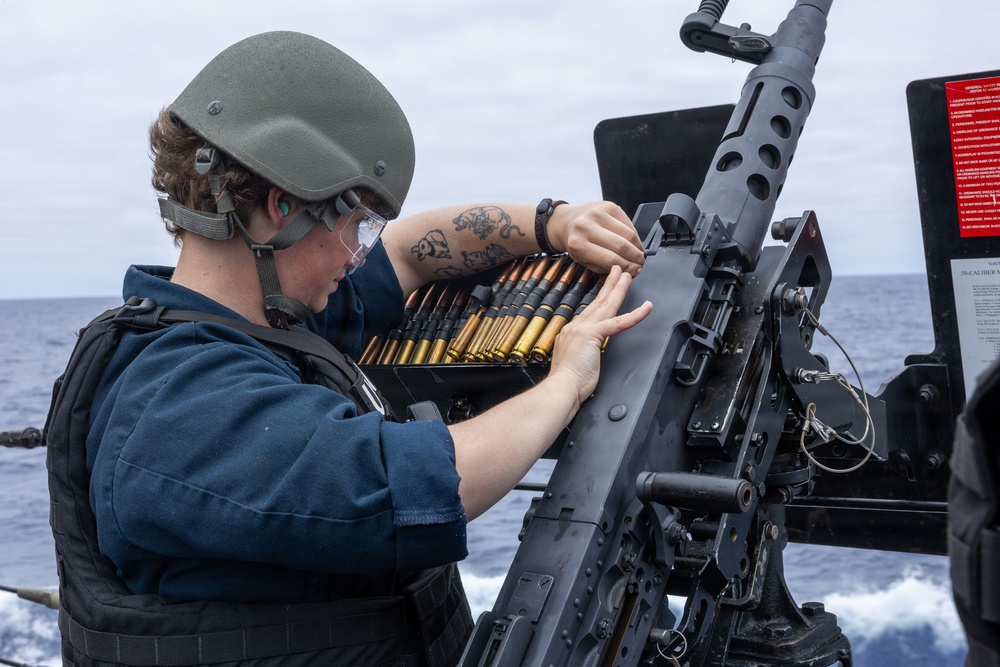 A military sailor in full protective gear is loading a belt-fed heavy machine gun mounted on the deck of a naval vessel at sea. The individual is wearing a combat helmet, ballistic eyewear, and a tactical vest while handling a linked ammunition belt. The setting appears to be an open ocean environment, suggesting active naval operations or weapons training.
