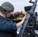 A military sailor in full protective gear is loading a belt-fed heavy machine gun mounted on the deck of a naval vessel at sea. The individual is wearing a combat helmet, ballistic eyewear, and a tactical vest while handling a linked ammunition belt. The setting appears to be an open ocean environment, suggesting active naval operations or weapons training.