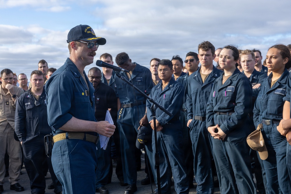A naval officer in dark blue coveralls and a ship's ball cap is addressing a large group of sailors at a microphone on what appears to be a ship's deck or outdoor area. The speaker holds papers and appears to be delivering a formal address or briefing to the assembled crew. The group of sailors, dressed in matching navy blue coveralls, stands attentively in formation.