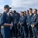 A naval officer in dark blue coveralls and a ship's ball cap is addressing a large group of sailors at a microphone on what appears to be a ship's deck or outdoor area. The speaker holds papers and appears to be delivering a formal address or briefing to the assembled crew. The group of sailors, dressed in matching navy blue coveralls, stands attentively in formation.