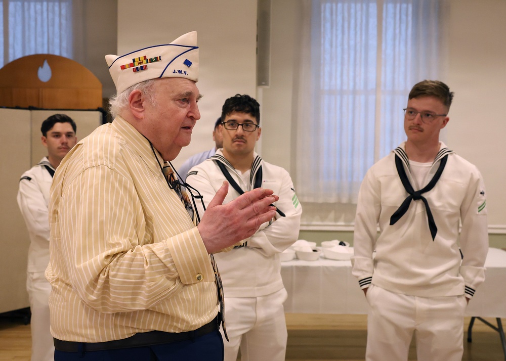 An elderly veteran wearing a white garrison cap with ribbon decorations and 'J.W.V.' (Jewish War Veterans) insignia is speaking to a group of young U.S. Navy sailors in white dress uniforms. The setting appears to be an indoor community or ceremonial hall. The veteran appears to be sharing stories or addressing the sailors in what looks like a formal or commemorative gathering.