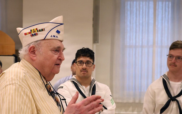 An elderly veteran wearing a white garrison cap with ribbon decorations and 'J.W.V.' (Jewish War Veterans) insignia is speaking to a group of young U.S. Navy sailors in white dress uniforms. The setting appears to be an indoor community or ceremonial hall. The veteran appears to be sharing stories or addressing the sailors in what looks like a formal or commemorative gathering.