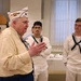 An elderly veteran wearing a white garrison cap with ribbon decorations and 'J.W.V.' (Jewish War Veterans) insignia is speaking to a group of young U.S. Navy sailors in white dress uniforms. The setting appears to be an indoor community or ceremonial hall. The veteran appears to be sharing stories or addressing the sailors in what looks like a formal or commemorative gathering.