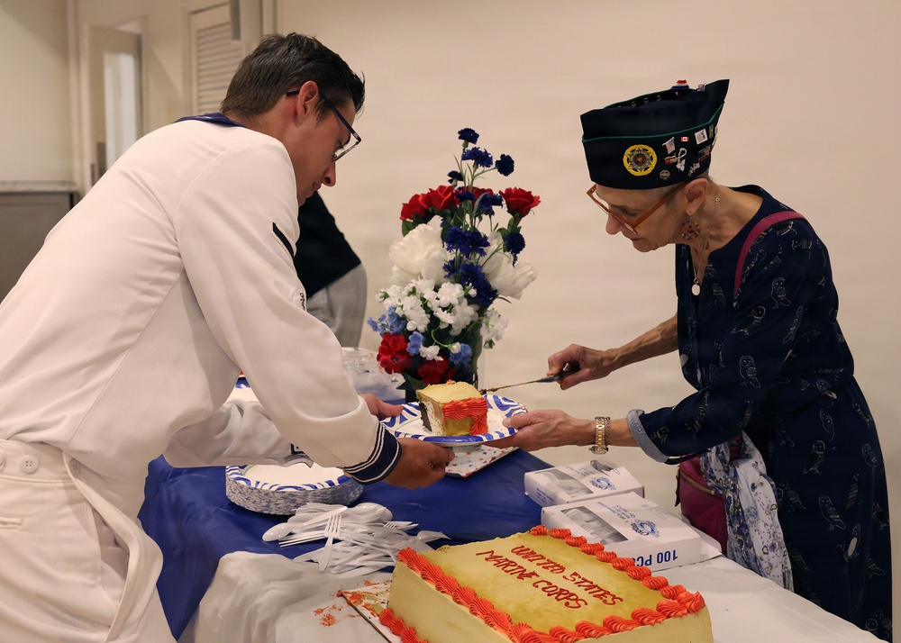 A cake-cutting ceremony appears to be taking place at a United States Marine Corps event. A person in a white uniform is serving cake to an elderly woman wearing a military-style garrison cap with insignia. The cake is decorated with red frosting and reads 'United States Marine Corps' on it.
