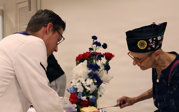 A cake-cutting ceremony appears to be taking place at a United States Marine Corps event. A person in a white uniform is serving cake to an elderly woman wearing a military-style garrison cap with insignia. The cake is decorated with red frosting and reads 'United States Marine Corps' on it.