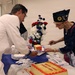 A cake-cutting ceremony appears to be taking place at a United States Marine Corps event. A person in a white uniform is serving cake to an elderly woman wearing a military-style garrison cap with insignia. The cake is decorated with red frosting and reads 'United States Marine Corps' on it.