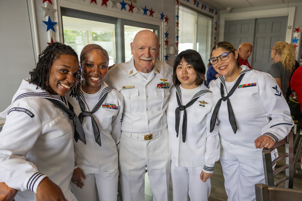 Five U.S. Navy personnel pose together for a group photo in what appears to be a decorated indoor venue. The group consists of one senior officer in dress whites with multiple ribbons and insignia, flanked by four junior enlisted sailors in standard white dress uniforms with black neckerchiefs. The setting is decorated with red, white, and blue star garlands, suggesting a patriotic or military celebration event.