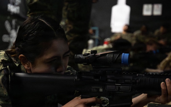 A female service member in digital camouflage uniform is in a prone firing position, aiming a rifle equipped with an optical scope during what appears to be an indoor marksmanship training exercise. Other personnel in similar uniforms are visible in the background, also in prone positions. A white silhouette target is visible downrange in the background.