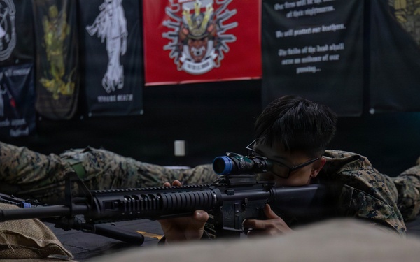 Two individuals in military camouflage uniforms (MARPAT pattern) are in the prone position aiming rifles with optical scopes, likely during a marksmanship training exercise. The setting appears to be an indoor range or training facility decorated with military-themed banners and flags on the wall behind them. The shooter in the foreground is actively looking through a rifle scope mounted on what appears to be an M16/AR-15 platform rifle.