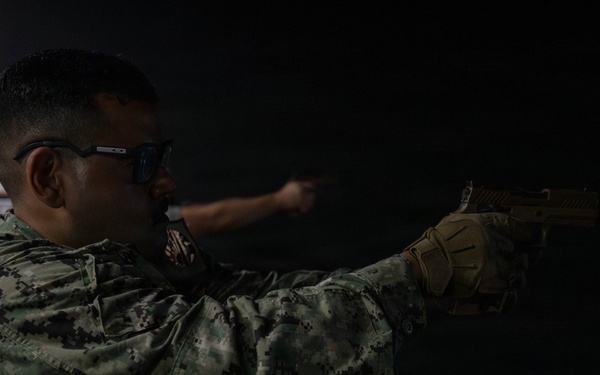 A military service member in digital camouflage uniform is practicing at an indoor shooting range. The individual is in a two-handed shooting stance, aiming a tan/FDE-colored pistol downrange toward a paper target. The setting appears to be a controlled indoor range environment with low ambient lighting.