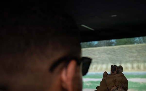 A military or law enforcement personnel in camouflage uniform is aiming a pistol at a projected shooting range simulation screen. The individual is wearing tactical gloves, sunglasses, and a watch, and appears to be inside a firearms training simulator. The projected screen displays a realistic outdoor shooting range with paper targets visible downrange.