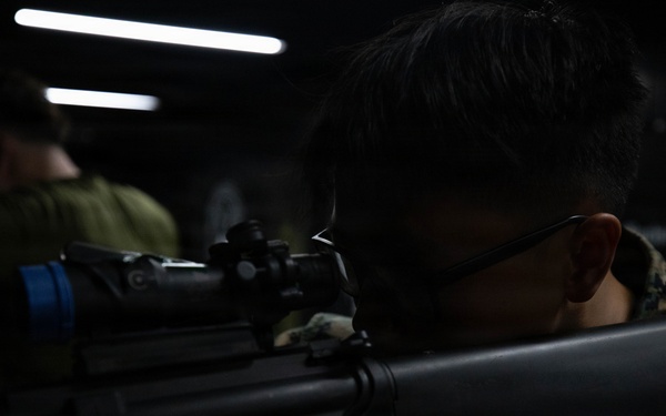 A military service member in digital camouflage uniform is aiming a rifle equipped with an optical scope in a low-light indoor environment. The shooter is in a proper firing stance with eye aligned to the optic. A second person in military attire is visible in the background.