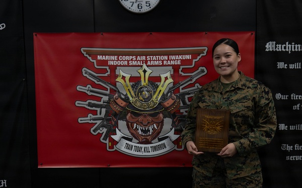 A U.S. Marine in woodland MARPAT camouflage uniform is posing with a wooden plaque award at Marine Corps Air Station Iwakuni's Indoor Small Arms Range. The Marine is smiling and standing in front of a red banner bearing the MCAS Iwakuni Indoor Small Arms Range logo with the motto 'Train Today, Kill Tomorrow.' The setting appears to be an official award or recognition ceremony.