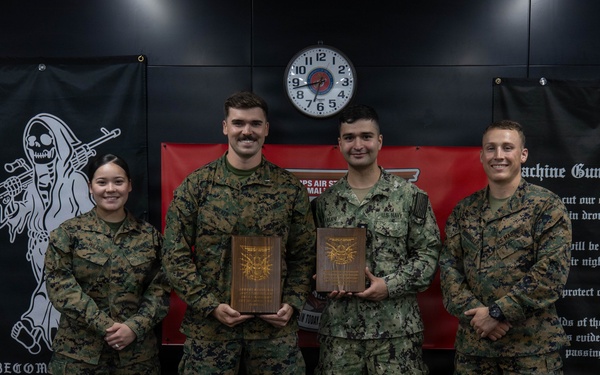 Four military personnel in camouflage uniforms pose for a formal group photo, with two individuals holding wooden plaques or awards. The setting appears to be an indoor military facility decorated with unit flags and a 'Machine Gunner's Creed' banner. Three individuals appear to be U.S. Marines in MARPAT camouflage, while one appears to be wearing Navy NWU Type III camouflage.