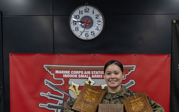 A U.S. Marine in MARPAT digital camouflage uniform stands holding three wooden plaques/trophies in front of a red banner reading 'Marine Corps Air Station Indoor Small Arms' with the motto 'Train Today, K[ill Tomorrow]'. The individual is smiling and appears to be posing for an award or recognition photo. A wall clock and electrical outlet are visible in the background.