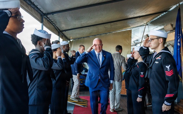 A civilian dignitary in a blue suit walks along a red carpet while U.S. Navy sailors in dress uniforms render salutes on both sides, forming a sideboy honor guard. The ceremony appears to be taking place on the deck of a naval vessel or pier under a canopy. An American flag and what appears to be a state flag are visible in the background.