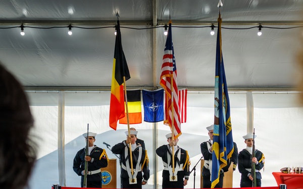 A formal military color guard ceremony is taking place inside a tent or temporary structure. Five U.S. Navy personnel in dress uniforms are standing at attention on a raised wooden platform, holding multiple flags including the U.S. flag, Belgian flag, NATO flag, and what appears to be a unit or command flag. Two audience members are visible in the foreground, observing the ceremony from behind a red rope barrier.