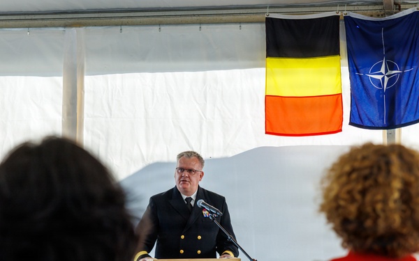 A military officer in dark naval dress uniform is delivering a speech at a formal ceremony from a wooden podium bearing an official seal. Three flags are displayed in the background: the Belgian flag, the NATO flag, and the United States flag. An audience is visible in the foreground, with attendees in civilian and formal attire.