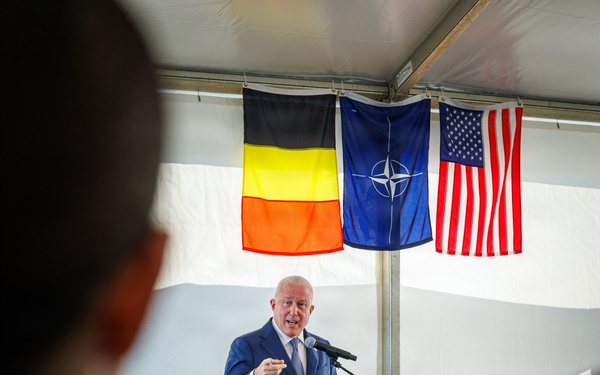 A man in a blue suit is delivering a speech at a wooden podium bearing what appears to be a U.S. Department of Defense or military seal. The event takes place under a tent with three flags displayed: the Belgian flag, the NATO flag, and the American flag. The scene suggests an official diplomatic or military ceremony involving the United States, NATO, and Belgium.