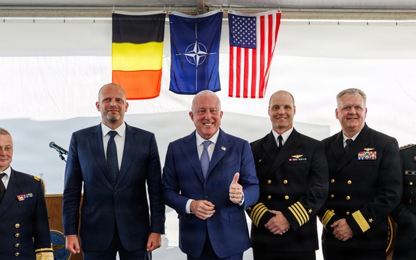Six individuals pose for a formal group photograph under a tent or covered venue. Three are in civilian business suits and three are in military uniforms representing different branches and nationalities. Behind them hang three flags: the Belgian flag, the NATO flag, and the United States flag.