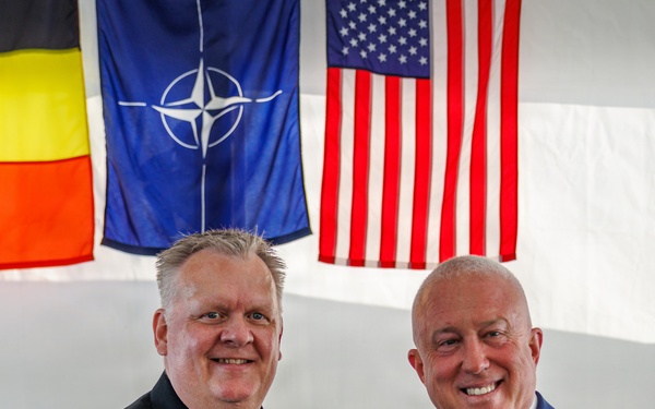 Two men are shaking hands in what appears to be an official military or diplomatic ceremony. One man is wearing a dark naval dress uniform with gold rank stripes, medals, and insignia, while the other is dressed in a civilian blue suit with an American flag lapel pin and is holding a blue velvet presentation case. The background displays three flags: the Belgian flag, the NATO flag, and the United States flag.