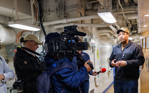 A media interview is taking place inside what appears to be a naval vessel or large ship, with a person in a dark jacket and cap speaking to a group of journalists and camera operators. A videographer with a professional broadcast camera and a reporter holding a red-tipped microphone are capturing the interview. Several other media personnel and observers are present in the corridor-like interior space.