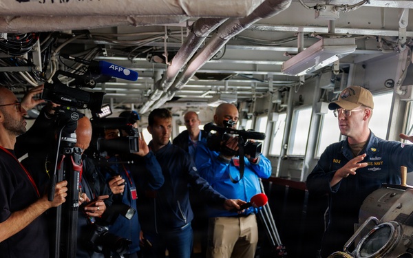 A U.S. Navy officer is conducting a press briefing on the bridge of a naval vessel, addressing a group of journalists and camera crews. The officer, wearing a tan cap and U.S. Navy uniform, is gesturing while speaking near the ship's helm controls. Multiple media organizations are present, including AFP, with cameras and microphones capturing the briefing.