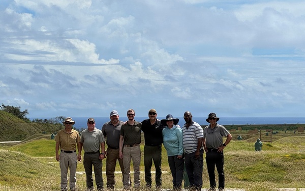 A group of eight adults poses together for a group photo on a grassy, undulating terrain that appears to be an outdoor shooting range or training facility. The ocean is visible in the background along with a partly cloudy sky. Several human-shaped targets or figures can be seen scattered across the range in the background.