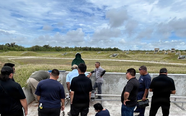 A group of approximately 10-12 individuals are gathered around a concrete shooting position or firing point structure at what appears to be an outdoor shooting range. The group appears to be receiving instruction or a demonstration, with one person crouching near equipment attached to the concrete barrier. The setting features multiple earthen berms and target positions visible in the background across a flat, grassy field.
