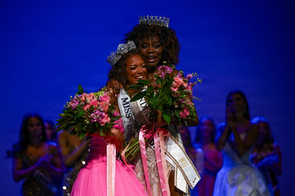 Two women are sharing an emotional embrace on what appears to be a pageant stage. Both are wearing crowns and sashes, with one in a pink gown and the other in a beaded/sequined gown, both holding pink floral bouquets. The setting suggests a beauty pageant crowning ceremony with other contestants visible in the background.