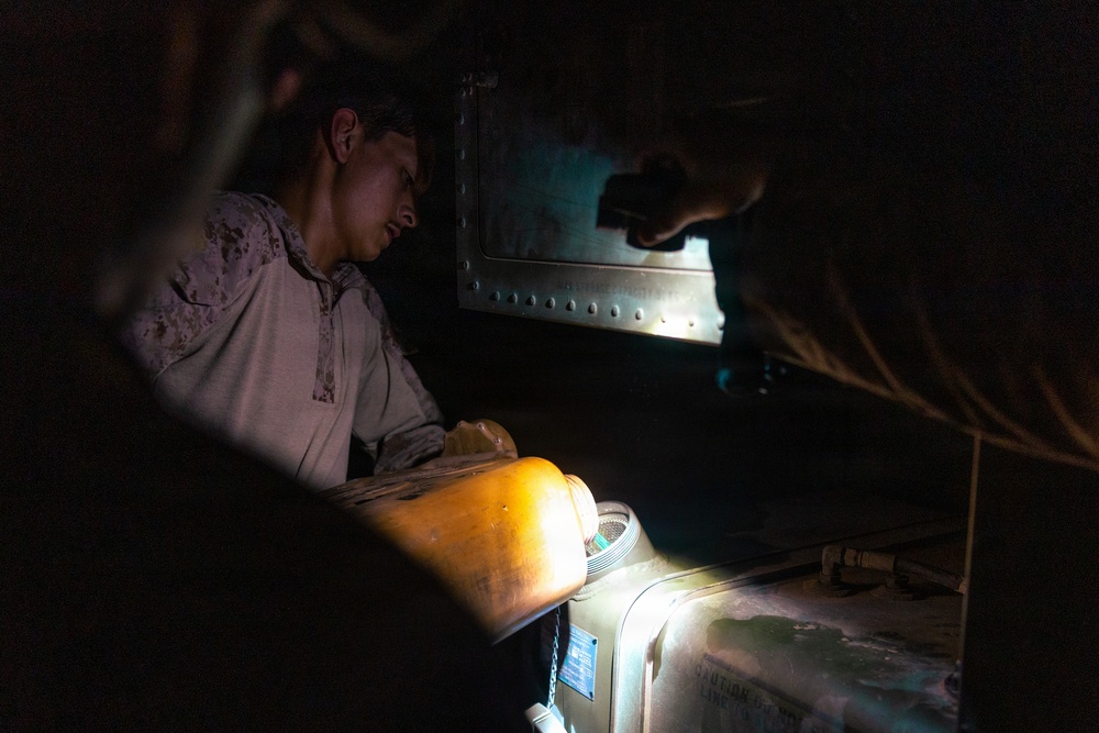A person wearing camouflage military-style clothing is working in a dark, confined space illuminated by a work light. They appear to be performing maintenance or inspection on mechanical or industrial equipment. A second individual is partially visible in the upper right, holding what appears to be a flashlight or tool.