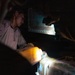 A person wearing camouflage military-style clothing is working in a dark, confined space illuminated by a work light. They appear to be performing maintenance or inspection on mechanical or industrial equipment. A second individual is partially visible in the upper right, holding what appears to be a flashlight or tool.