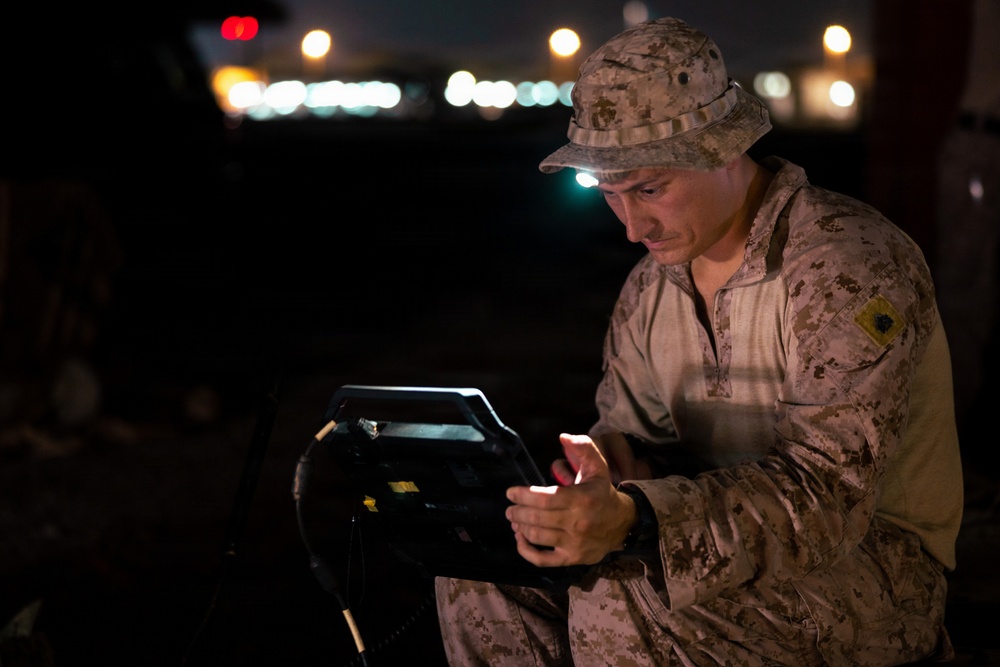 A military service member in desert camouflage (MARPAT pattern) operates a ruggedized electronic device or control unit at night. The soldier is wearing a boonie hat with an attached headlamp that illuminates his face and the equipment. The background shows blurred lights consistent with a base or airfield environment.