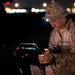 A military service member in desert camouflage (MARPAT pattern) operates a ruggedized electronic device or control unit at night. The soldier is wearing a boonie hat with an attached headlamp that illuminates his face and the equipment. The background shows blurred lights consistent with a base or airfield environment.