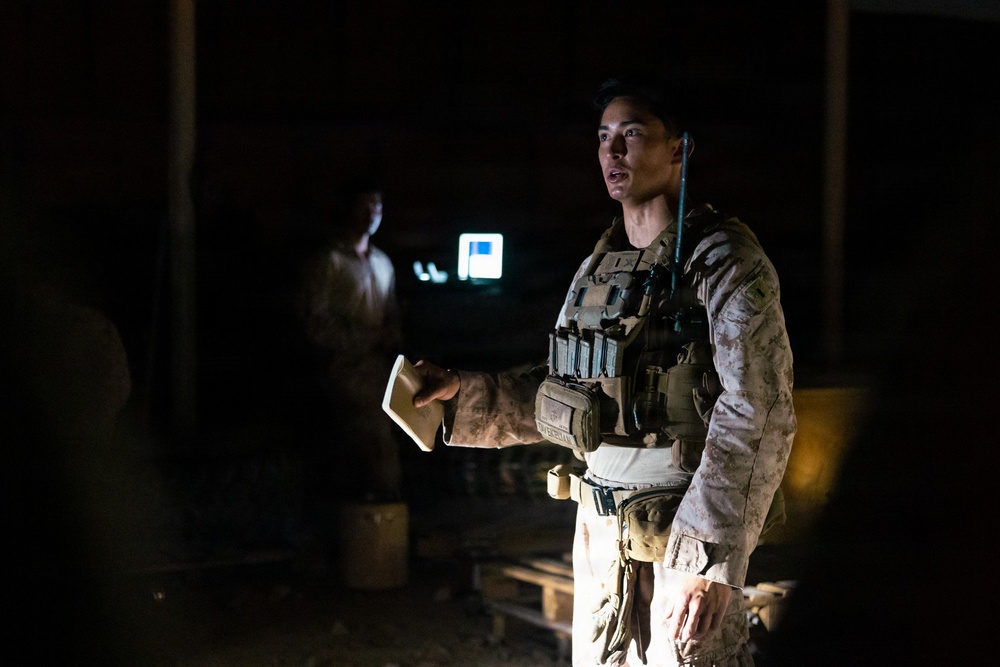 A military service member in desert camouflage uniform and tactical vest is standing in a dimly lit indoor or covered outdoor space at night. The individual appears to be holding a small notebook or card and may be briefing or communicating with others. A second figure is visible in the dark background near a glowing screen or light source.