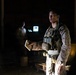A military service member in desert camouflage uniform and tactical vest is standing in a dimly lit indoor or covered outdoor space at night. The individual appears to be holding a small notebook or card and may be briefing or communicating with others. A second figure is visible in the dark background near a glowing screen or light source.
