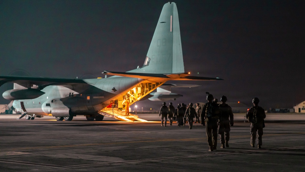 A nighttime military operation showing armed personnel boarding a large military transport aircraft via its rear ramp. The aircraft, consistent with a C-130 Hercules configuration, is illuminated from within, casting warm light onto the tarmac. Multiple soldiers in full combat gear are walking toward and up the lowered cargo ramp.