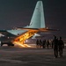 A nighttime military operation showing armed personnel boarding a large military transport aircraft via its rear ramp. The aircraft, consistent with a C-130 Hercules configuration, is illuminated from within, casting warm light onto the tarmac. Multiple soldiers in full combat gear are walking toward and up the lowered cargo ramp.