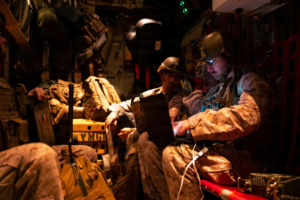 Military personnel, appearing to be U.S. Marines based on their MARPAT desert camouflage uniforms, are seated inside a military aircraft during what appears to be a nighttime operation. The soldiers are equipped with tactical gear, helmets, and night-vision devices, and are reviewing information on a tablet or electronic device. The interior is dimly lit with warm amber lighting, suggesting a low-light operational environment.