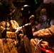 Military personnel, appearing to be U.S. Marines based on their MARPAT desert camouflage uniforms, are seated inside a military aircraft during what appears to be a nighttime operation. The soldiers are equipped with tactical gear, helmets, and night-vision devices, and are reviewing information on a tablet or electronic device. The interior is dimly lit with warm amber lighting, suggesting a low-light operational environment.