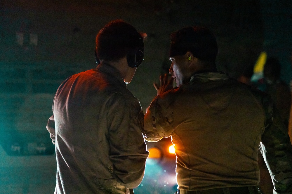Two individuals in military-style uniforms are engaged in close conversation in a dimly lit environment. One person is wearing over-ear headphones or communication headset, while the other appears to have ear protection (yellow earplugs visible). The scene is illuminated by warm orange and cool teal ambient lighting, suggesting a nighttime operational or industrial setting.