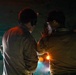 Two individuals in military-style uniforms are engaged in close conversation in a dimly lit environment. One person is wearing over-ear headphones or communication headset, while the other appears to have ear protection (yellow earplugs visible). The scene is illuminated by warm orange and cool teal ambient lighting, suggesting a nighttime operational or industrial setting.