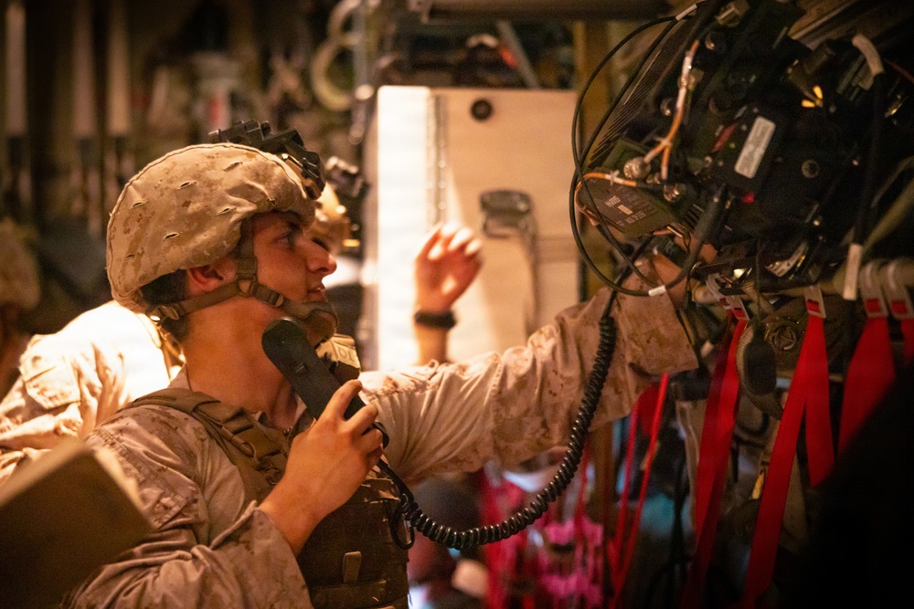 A U.S. Marine in desert MARPAT camouflage and combat helmet is operating communications equipment inside what appears to be a military aircraft or vehicle. The Marine is holding a handset/radio receiver to their mouth while reaching toward a complex panel of mounted electronic equipment. The interior setting suggests this is a command, control, or communications operation during a training exercise or mission.