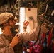 A U.S. Marine in desert MARPAT camouflage and combat helmet is operating communications equipment inside what appears to be a military aircraft or vehicle. The Marine is holding a handset/radio receiver to their mouth while reaching toward a complex panel of mounted electronic equipment. The interior setting suggests this is a command, control, or communications operation during a training exercise or mission.