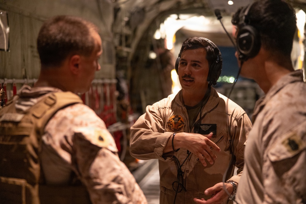 Three military personnel are engaged in conversation inside what appears to be a large military transport aircraft, likely a C-17 or C-130. The central figure is wearing a tan flight suit with a squadron patch and aviation wings, wearing a communications headset. The other two individuals are wearing desert camouflage uniforms with tactical vests.