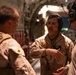 Three military personnel are engaged in conversation inside what appears to be a large military transport aircraft, likely a C-17 or C-130. The central figure is wearing a tan flight suit with a squadron patch and aviation wings, wearing a communications headset. The other two individuals are wearing desert camouflage uniforms with tactical vests.