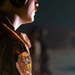 A close-up shot of a figure wearing a worn, tan/brown jacket or flight suit with a prominent embroidered patch on the shoulder. The patch features what appears to be a bear or wolf-like creature in golden-orange tones with text on a banner beneath it. The figure also wears goggles or headgear pushed up on their head, suggesting an aviator or adventurer aesthetic.