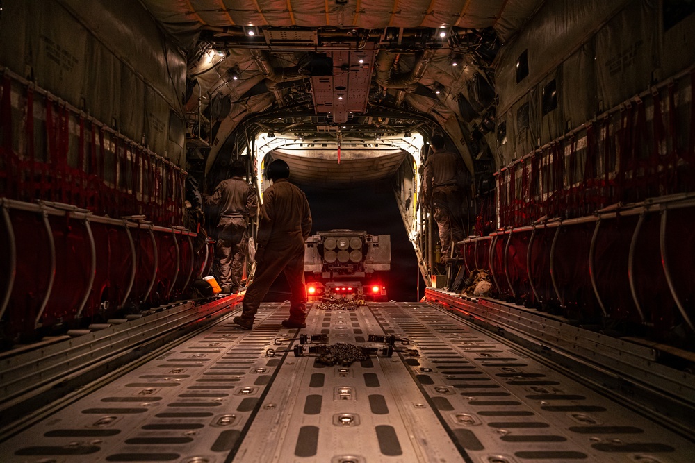 The image shows the interior of a large military cargo aircraft, likely a C-17 Globemaster or C-5 Galaxy, with the rear ramp open. Personnel in military flight suits are overseeing the loading or unloading of a military vehicle or weapons system via the rear cargo ramp at night. The cargo hold is illuminated by interior lighting, with red tail lights from the vehicle visible near the ramp opening.